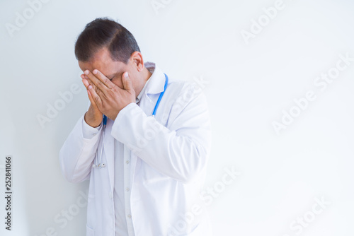 Middle age doctor man wearing stethoscope and medical coat over white background with sad expression covering face with hands while crying. Depression concept.