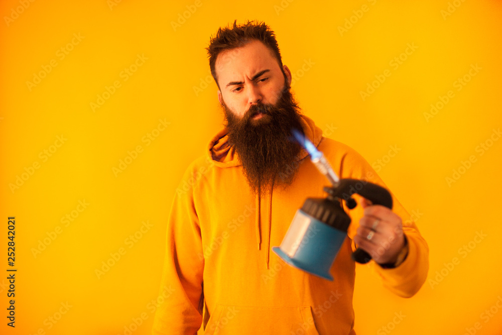 Bearded handy man holding blow torch over yellow background Bearded handy man holding blow torch over yellow background
