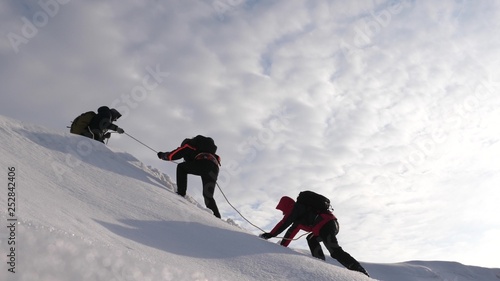 Travelers climb rope to their victory through snow uphill in strong wind. tourists in winter work together as team overcoming difficulties. three Alpenists in winter climb rope on mountain.
