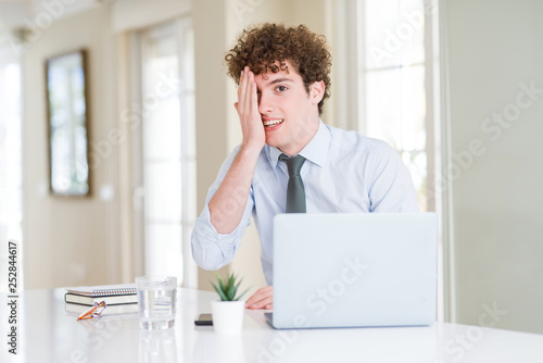 Young business man working with computer laptop at the office covering one eye with hand with confident smile on face and surprise emotion.