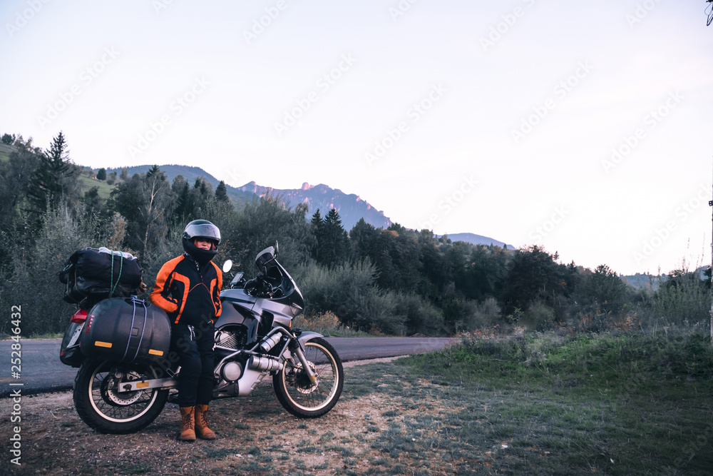 woman biker with adventure motorcycle parking on the side road, sunset, Ceahlau mountains and forest, Romania, tourism travel concept, motorcyclists way, copy space