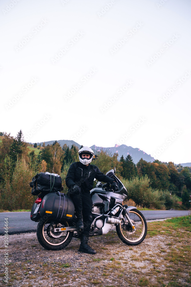 Rider Man and off tourism adventure motorcycles with side bags and equipment for long road trip, travel touring concept, Ceahlau, Romania, mountains on background, sunset evening, vertical photo