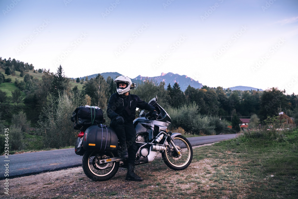 Rider Man and off tourism adventure motorcycles with side bags and equipment for long road trip, travel touring concept, Ceahlau, Romania, mountains on background, sunset evening