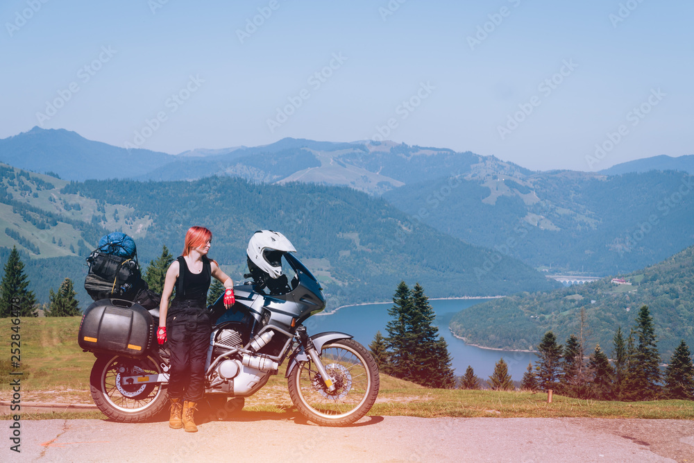 woman biker with adventure motorcycle on road, Ceahlau mountains, Romania, tourism travel concept, motorcyclists way, copy space, lake Lzvorul Muntelui, beautiful sunny day