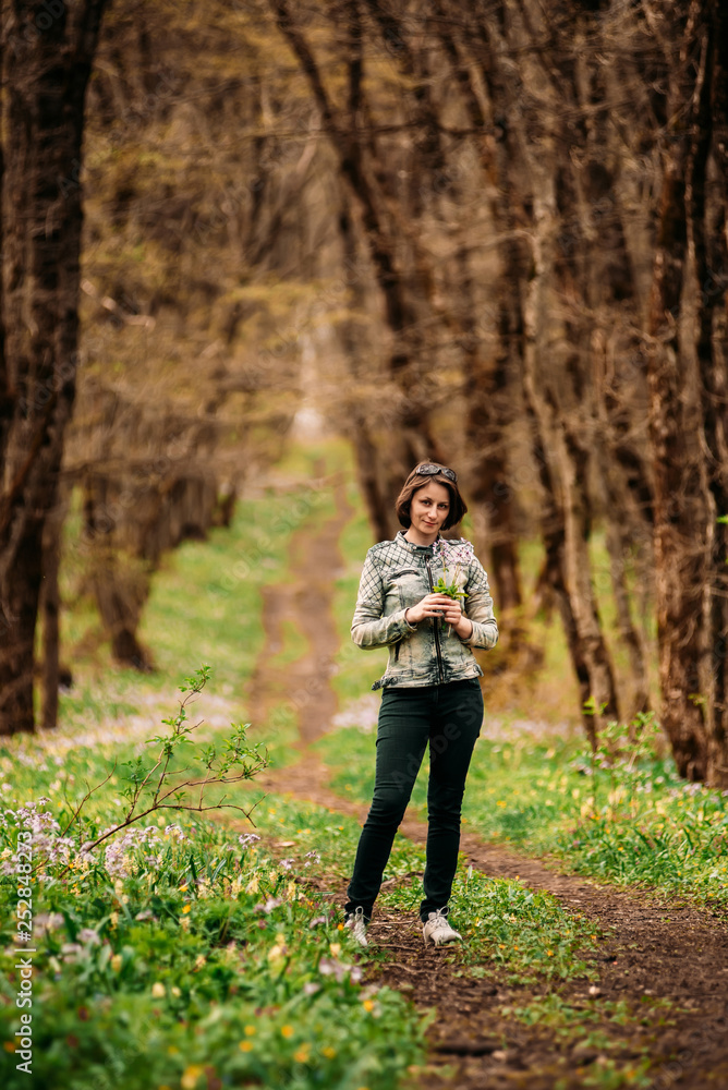 Photo of happy middle-aged woman in jacket standing in spring forest with flower in hand
