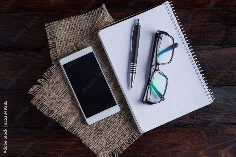 White coffee cup with notepad, glasses, pen and mobile phone on a dark wooden background and linen fabric White coffee cup with notepad, glasses, pen and mobile phone on a dark wooden background and linen fabric