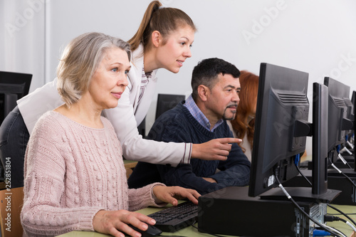 Young female teacher helping mature people to use computer