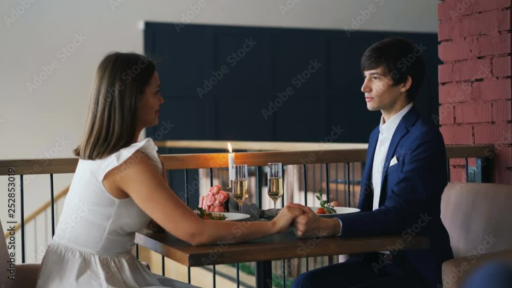 Beautiful couple is having romantic date in restaurant when young man is making proposal to happy woman giving her ring and kissing her hand, girl is laughing. Beautiful couple is having romantic date in restaurant when young man is making proposal to happy woman giving her ring and kissing her hand, girl is laughing.