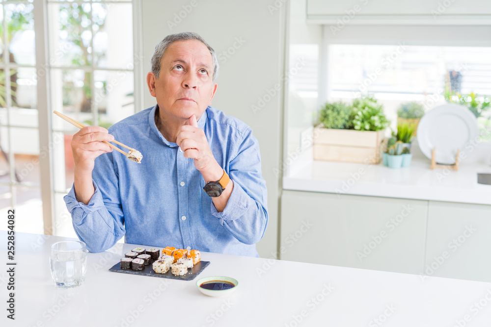 Handsome senior man eating take away sushi using chopsticks at home serious face thinking about question, very confused idea Handsome senior man eating take away sushi using chopsticks at home serious face thinking about question, very confused idea
