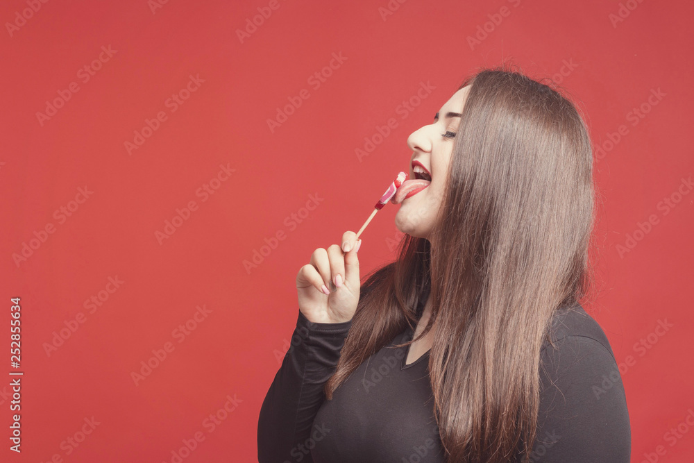 Obraz premium Model plus size with sweet donut, happy girl holding in hand candy and lick sweets, in black bodysuit. XXL female on red background