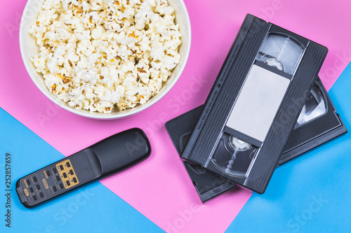 White bowl of popcorn, 2 VHS tapes and old fashioned remote control on a bright pink and blue background. Movie/ film industry. Entertainment. Unhealthy food/ snacks.