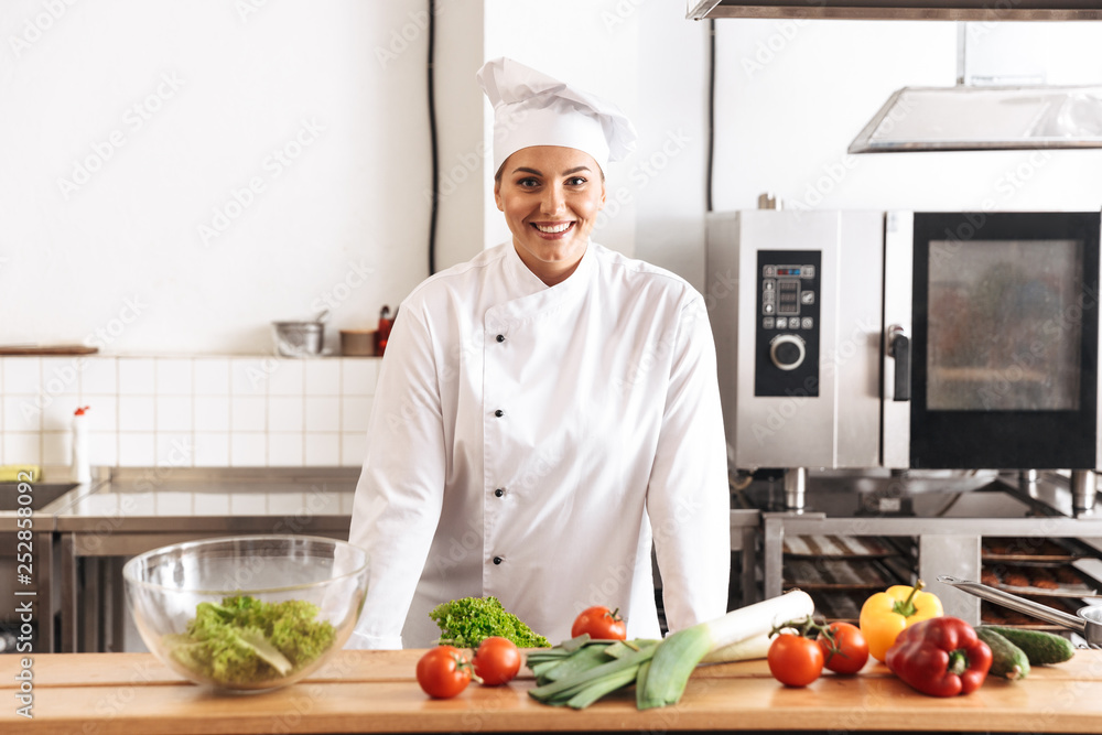 Photo of adult woman chef wearing white uniform cooking meal with fresh vegetables, in kitchen at the restaurant Photo of adult woman chef wearing white uniform cooking meal with fresh vegetables, in kitchen at the restaurant