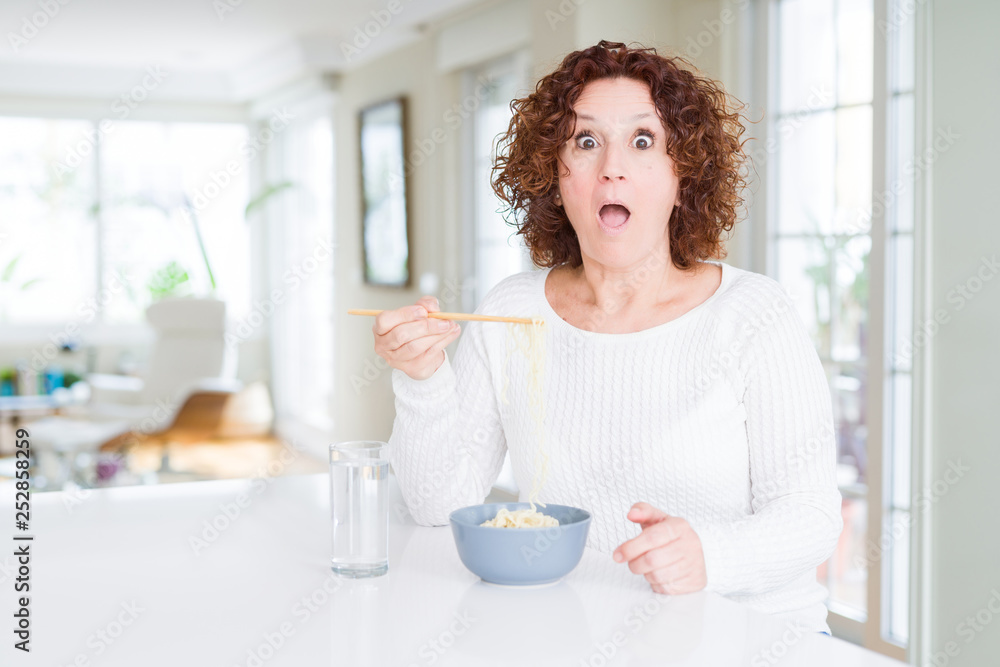 Senior woman eating asian noodles using chopsticks scared in shock with a surprise face, afraid and excited with fear expression Senior woman eating asian noodles using chopsticks scared in shock with a surprise face, afraid and excited with fear expression