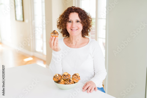 Senior woman eating chocolate chips muffins with a happy face standing and smiling with a confident smile showing teeth
