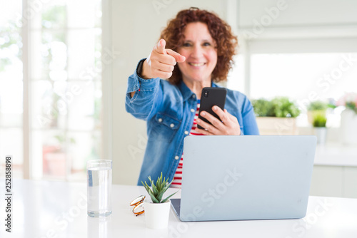 Senior woman using laptop and smartphone pointing with finger to the camera and to you, hand sign, positive and confident gesture from the front