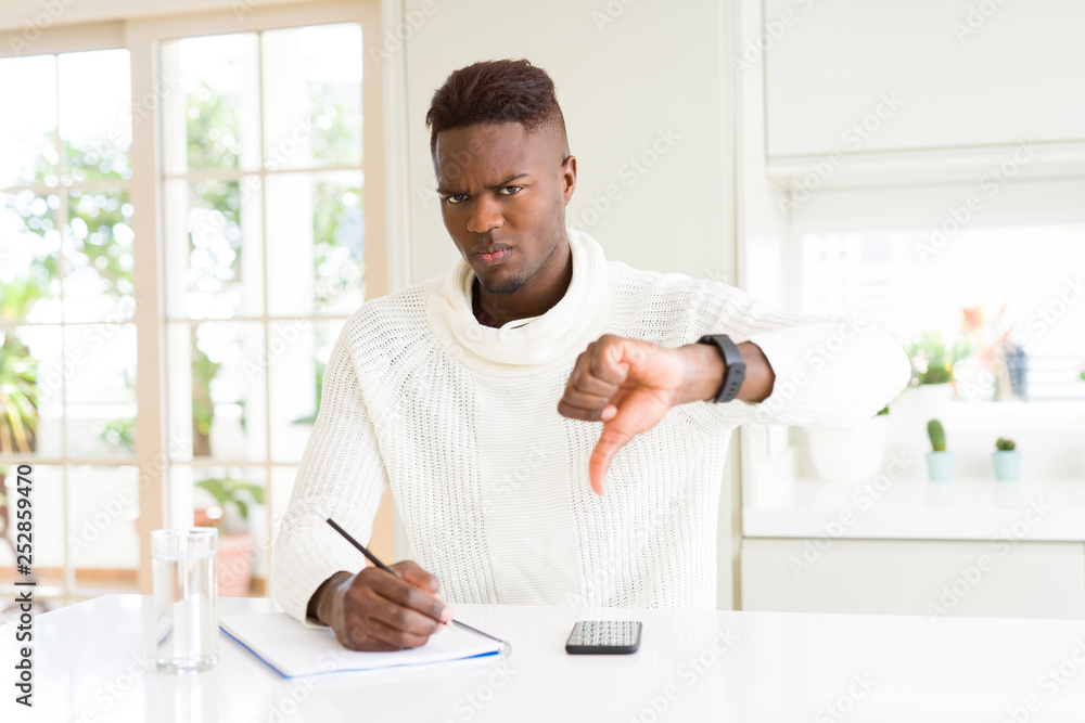 African american student man writing on a paper using a pencil with angry face, negative sign showing dislike with thumbs down, rejection concept