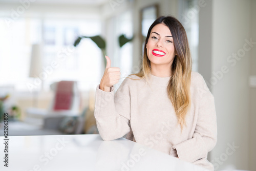Young beautiful woman wearing winter sweater at home doing happy thumbs up gesture with hand. Approving expression looking at the camera showing success.