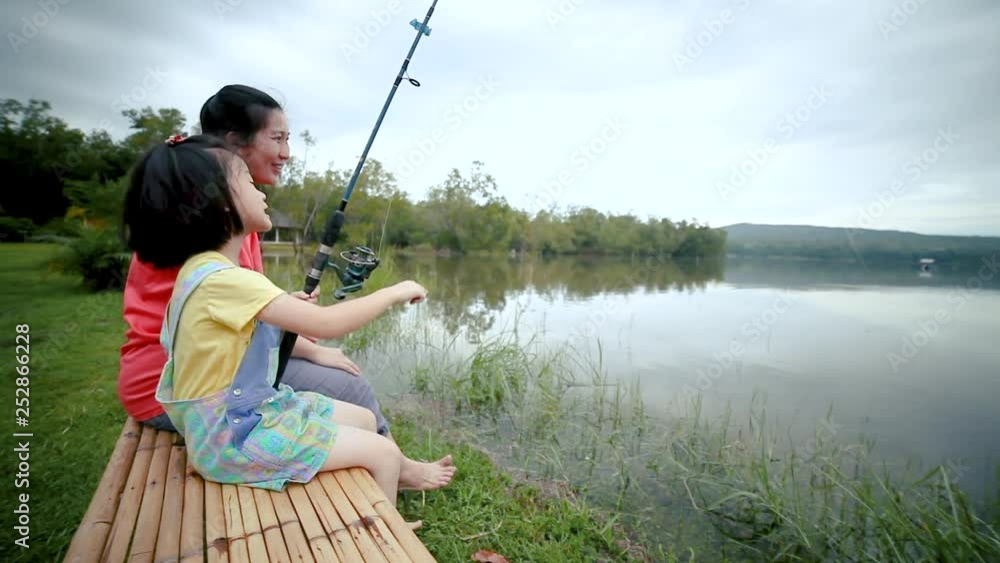Asian mother talking to her daughter While fishing, the couple sits on the banks of a tranquil evening lagoon.The girl is holding a fishing rod happily. Asian mother talking to her daughter While fishing, the couple sits on the banks of a tranquil evening lagoon.The girl is holding a fishing rod happily.