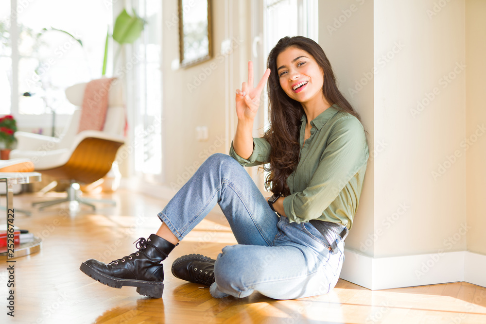 Young beautiful woman sitting on the floor at home smiling with happy face winking at the camera doing victory sign. Number two.