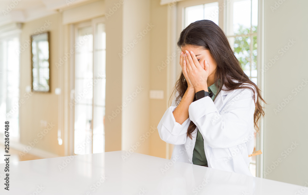Young woman wearing medical coat at the clinic as therapist or doctor with sad expression covering face with hands while crying. Depression concept. Young woman wearing medical coat at the clinic as therapist or doctor with sad expression covering face with hands while crying. Depression concept.