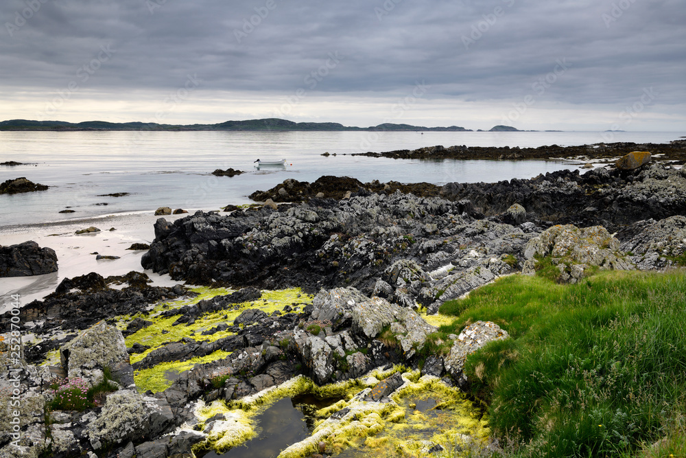 Sand beach and rocky shore with grass and algae on Isle of Iona with boat on Sound of Iona Inner Hebrides Scotland UK