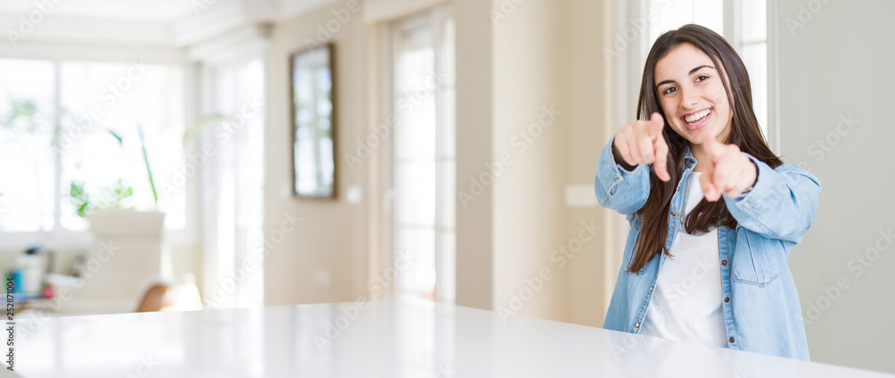Wide angle picture of beautiful young woman sitting on white table at home Pointing to you and the camera with fingers, smiling positive and cheerful Wide angle picture of beautiful young woman sitting on white table at home Pointing to you and the camera with fingers, smiling positive and cheerful