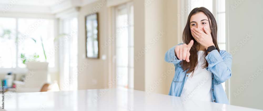 Wide angle picture of beautiful young woman sitting on white table at home Laughing of you, pointing to the camera with finger hand over mouth, shame expression Wide angle picture of beautiful young woman sitting on white table at home Laughing of you, pointing to the camera with finger hand over mouth, shame expression
