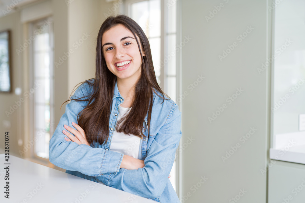 Beautiful young woman sitting on white table at home happy face smiling with crossed arms looking at the camera. Positive person.
