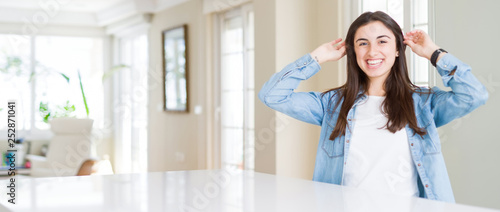 Wide angle picture of beautiful young woman sitting on white table at home Smiling pulling ears with fingers, funny gesture. Audition problem