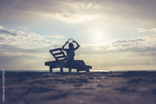 Girl in headphones is sitting on the sun lounger listening to music on the beach at sunset. Summer concept.