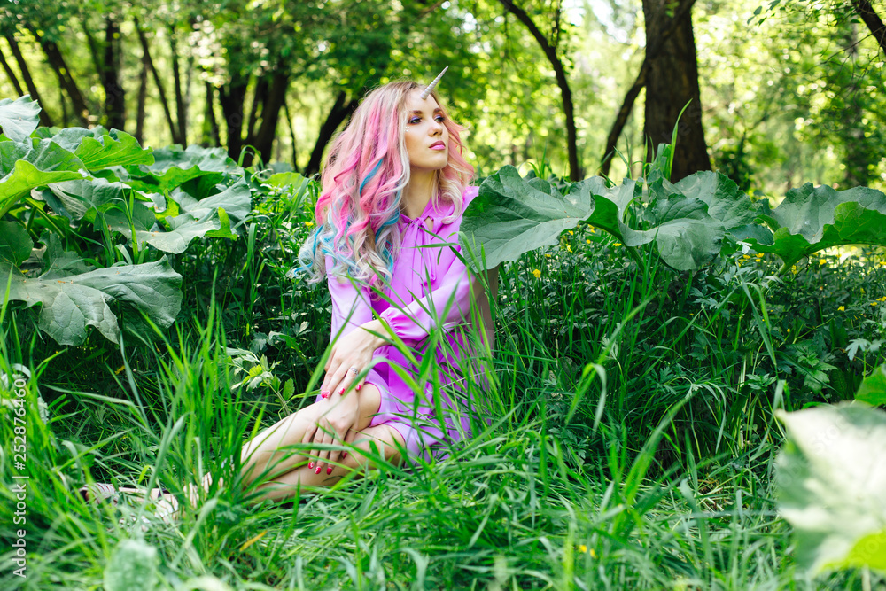 Fairy girl unicorn with rainbow hair and shiny makeup sitting on the grass. Fairy girl unicorn with rainbow hair and shiny makeup sitting on the grass.