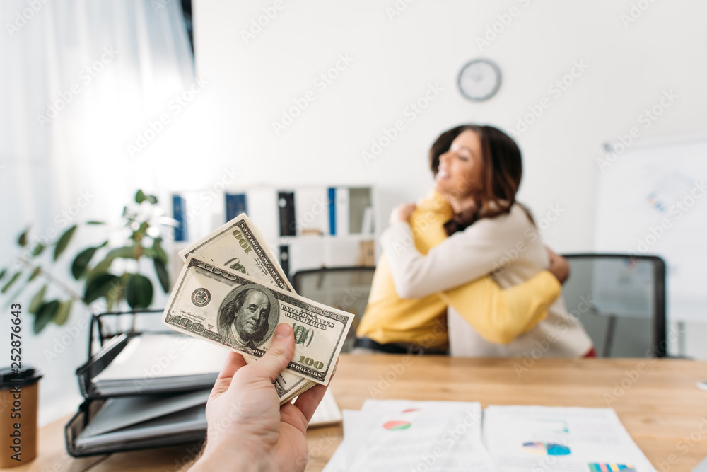selective focus of advisor at table holding dollar banknotes wile investors hugging in office