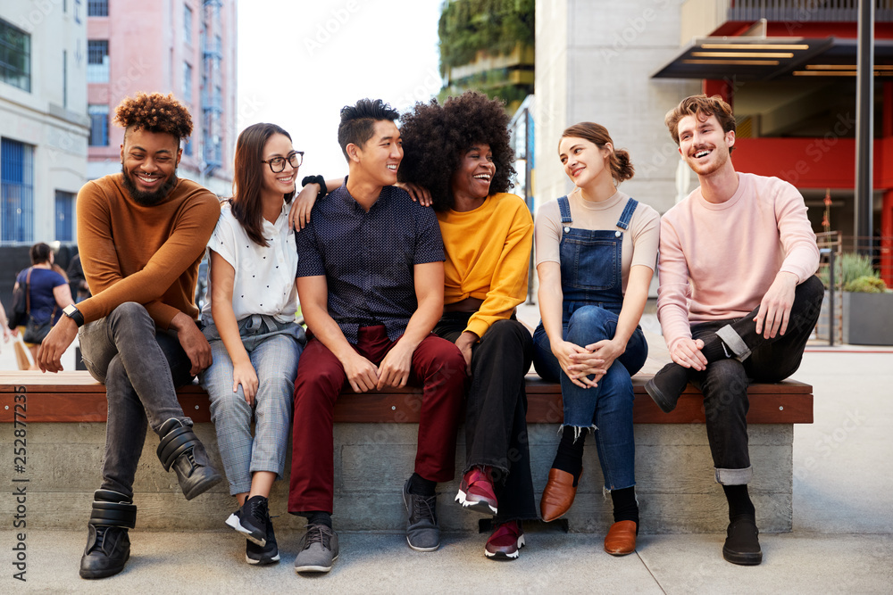 Fototapeta premium Six young adult friends sitting in a row on a bench in the street looking at each other, full length