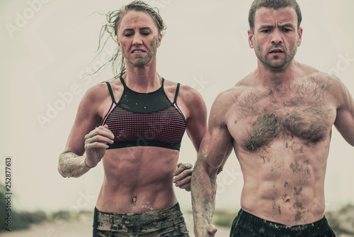 Muscular male and female athlete covered in mud running down a rough terrain with a desert background in an extreme sport race with grungy textured finish © Paul