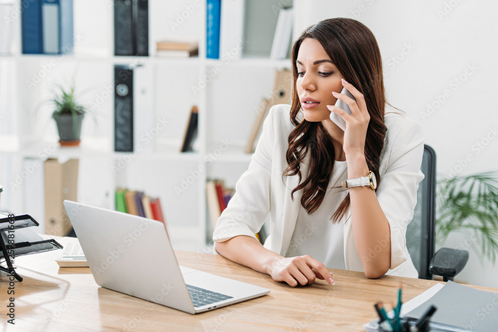 businesswoman sitting at table, looking to laptop and talking on smartphone in office businesswoman sitting at table, looking to laptop and talking on smartphone in office