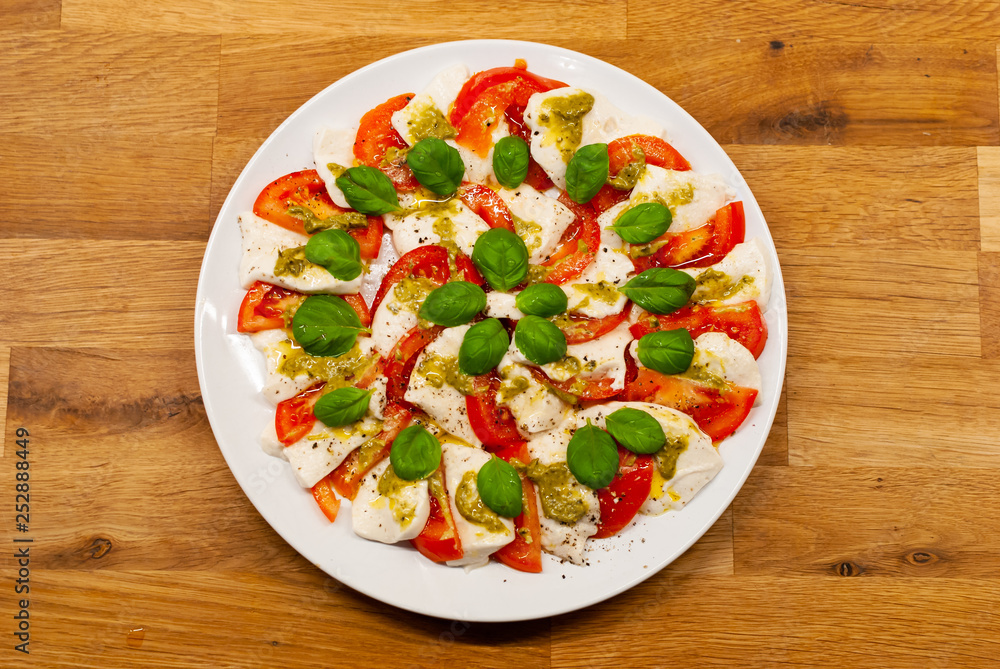 Top view of plate full of vegan Mozzarella made of cashew nuts with tomato slices, basil and pesto on a wooden table