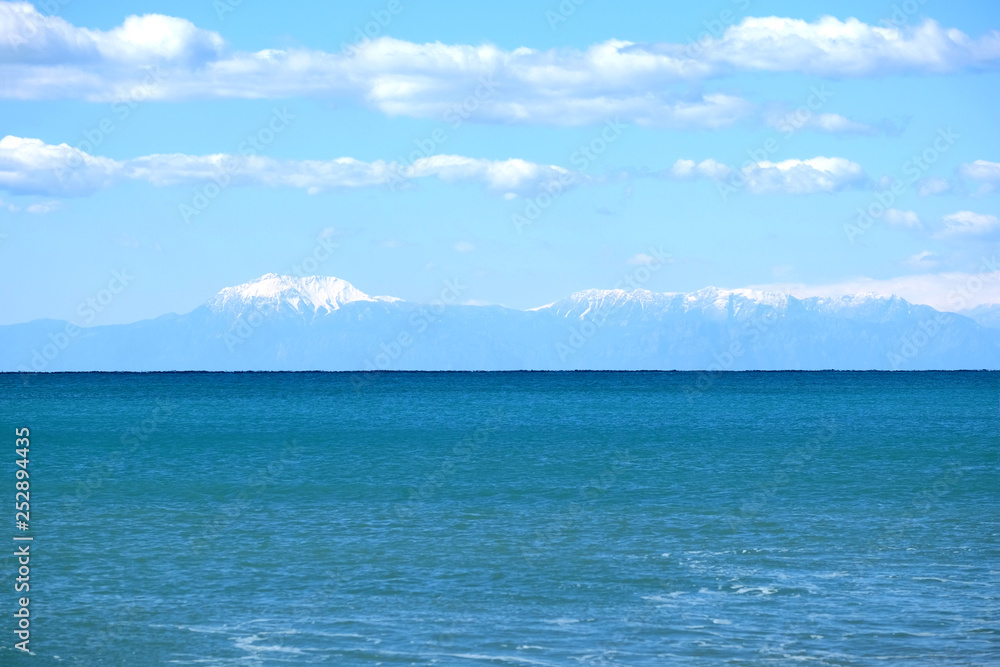 High mountains with white snow caps on tops far away on horizon in light haze after the sea under blue sky with cumulus clouds