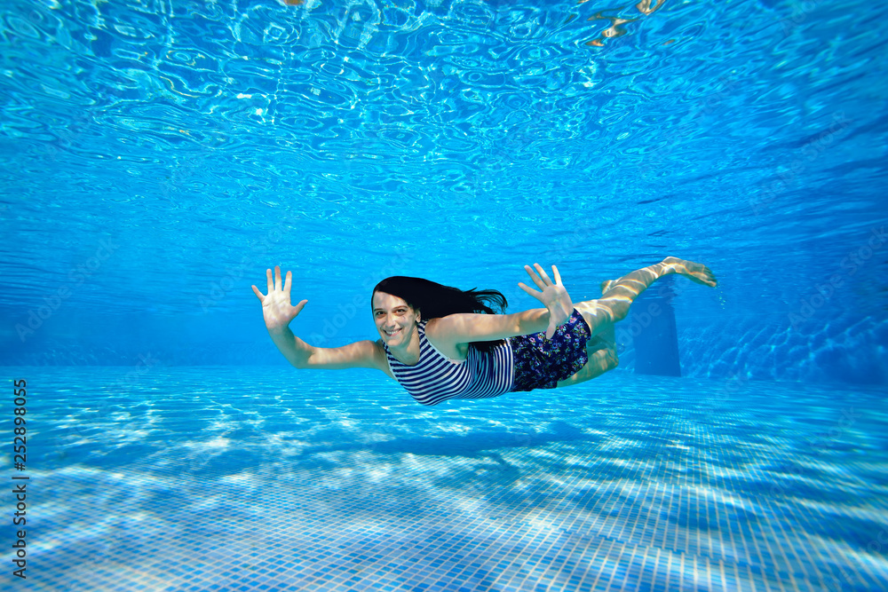 A cheerful sports girl swims under the water near the bottom of the pool in a striped swimsuit on a bright Sunny day, looks at the camera, waves her hands and smiles. Portrait. Underwater photography