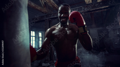 Hands of boxer over dark gym background. Strength, attack and motion concept. Fit african american model in movement. Naked muscular athlete in red gloves. Sporty man during boxing © master1305