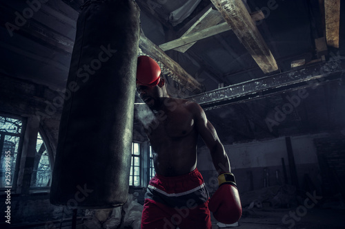 Hands of boxer over dark gym background. Strength, attack and motion concept. Fit african american model in movement. Naked muscular athlete in red gloves. Sporty man during boxing