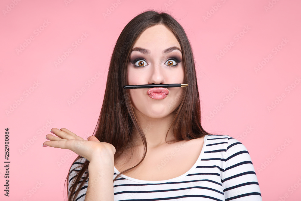 Young woman holding pen between nose and lips on pink background Young woman holding pen between nose and lips on pink background