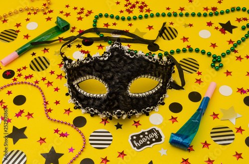 Top view decoration for party. Venice eye mask, plastic necklaces, party blowers and circles and stars confetti on yellow background