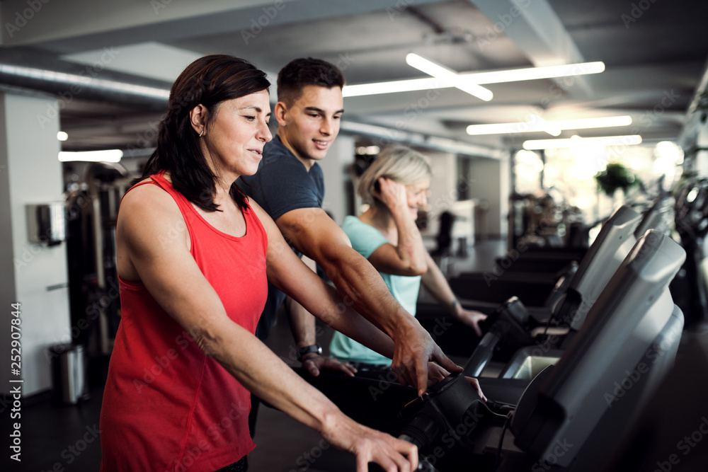 Two cheerful female seniors in gym with a young trainer doing cardio work out.