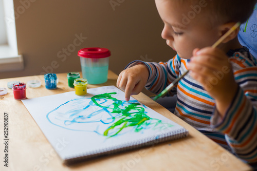 Left hand toddler drawing brush with gouache paint on paper in an album with several colorful brushes near it.