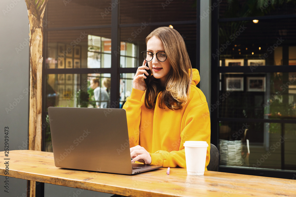 Girl in yellow hoodie sitting in cafe at table, talking on phone while looking to net-book screen. Woman works on laptop Girl in yellow hoodie sitting in cafe at table, talking on phone while looking to net-book screen. Woman works on laptop