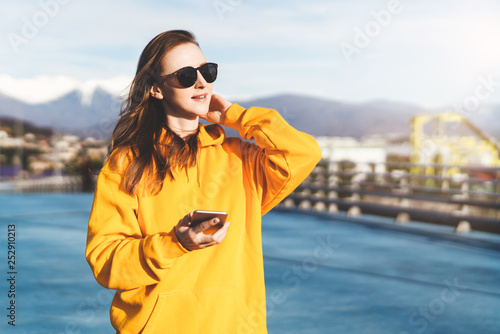 Front view. Girl in yellow hoodie and sunglasses holding smartphone while standing outdoors. In background mountains. © foxyburrow