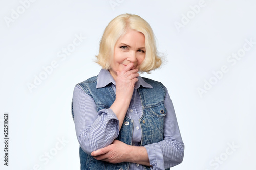 Thinking blonde senior woman looking at camera holding hand near mouth.