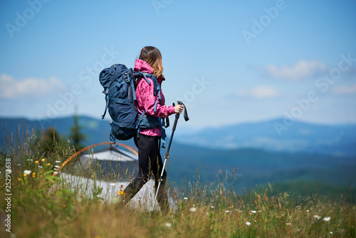 Back view of sporty girl traveller with backpack and trekking sticks near tent, wearing sports wear, hiking against blue sky and, enjoying sunny morning in the mountains. Camping lifestyle concept