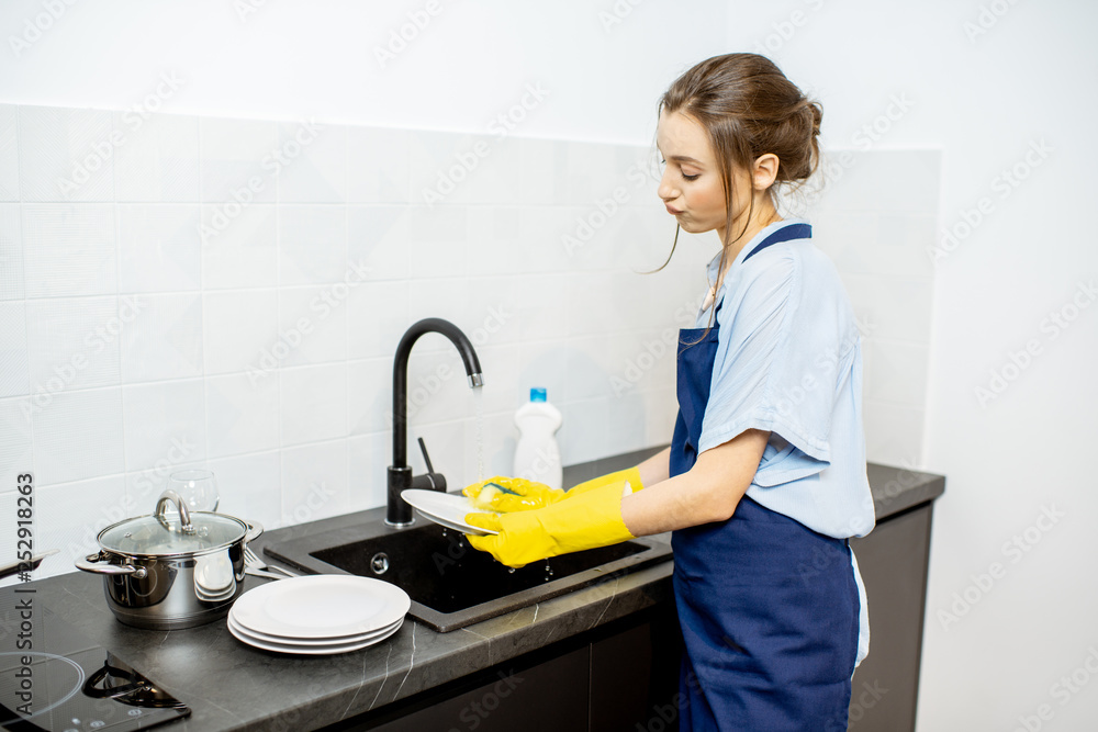 Young woman in apron feeling bored while washing dishes on the kitchen at home
