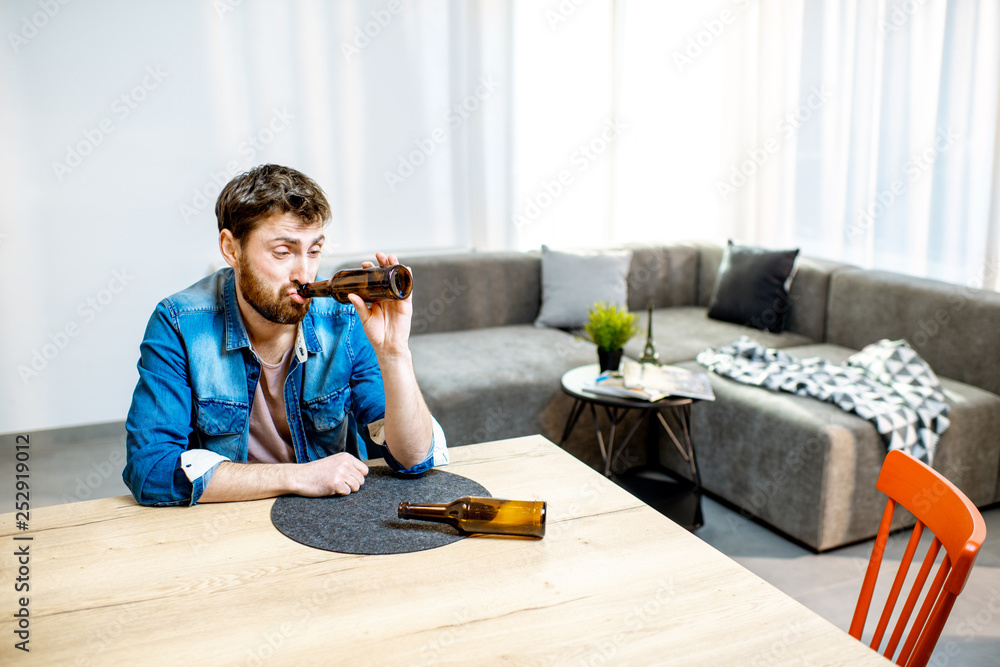 Drunk male alcoholic sitting with bottles feeling depressed and suffering from head ache at home Drunk male alcoholic sitting with bottles feeling depressed and suffering from head ache at home